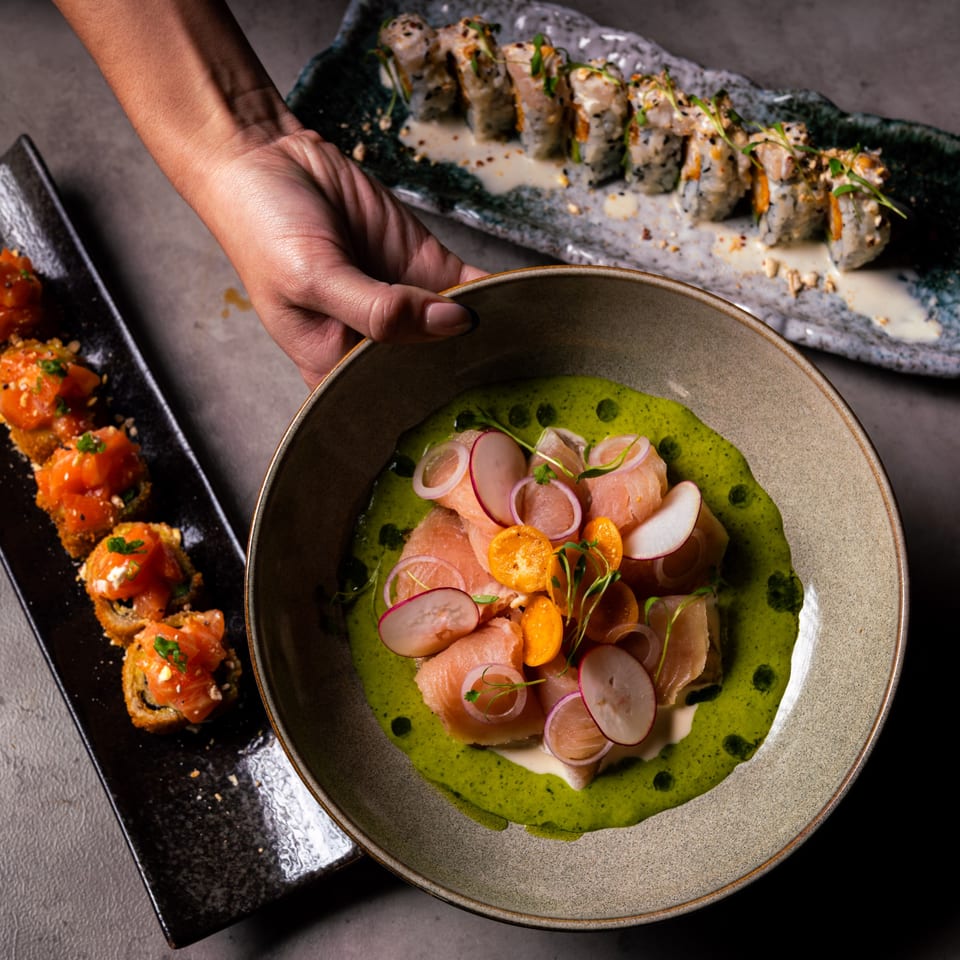 Hand holding a bowl of salmon ceviche with green sauce, alongside sushi rolls.