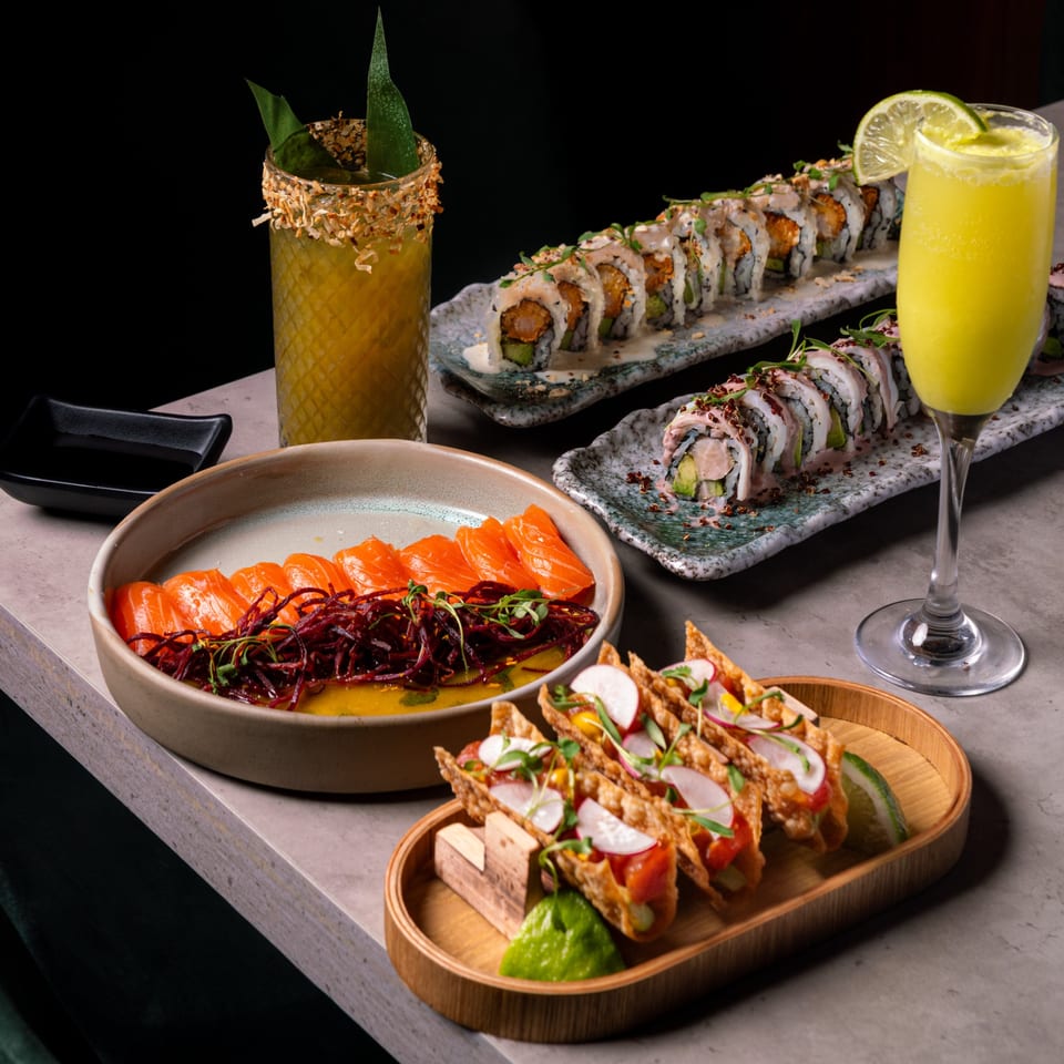 A vibrant spread of Sushi Jungle's sushi rolls, salmon sashimi, tacos, and two cocktails on a table.