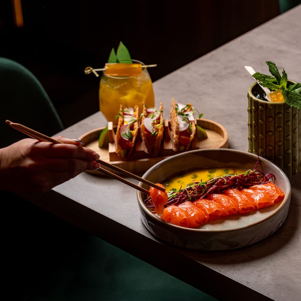 A hand uses chopsticks to pick up salmon sashimi from a plate, with tacos and cocktails nearby.
