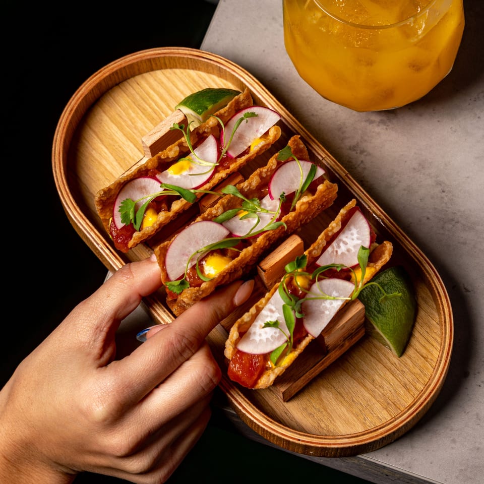 A hand reaches for a crispy sushi taco with tuna, radish, and microgreens on a wooden tray.